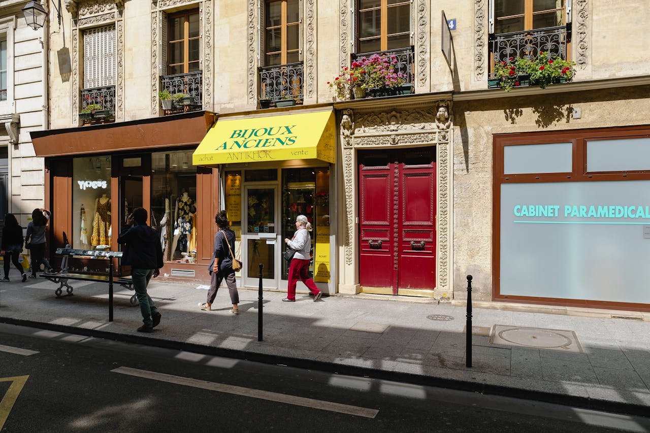 People walking by charming Parisian boutiques on a sunny day, showcasing vintage architecture and lively street life.