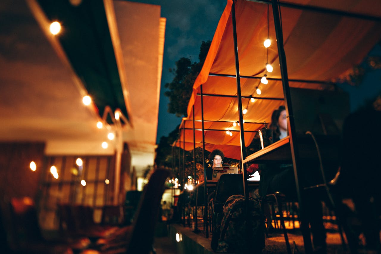 A vibrant evening cafe scene with people working on laptops under illuminated awnings.